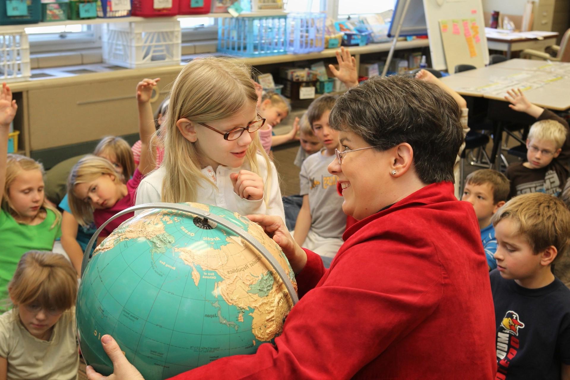 一位女世界语言老师向一个小女孩指出地球仪上的一个国家，而班上的其他人则坐在他们周围的地板上.