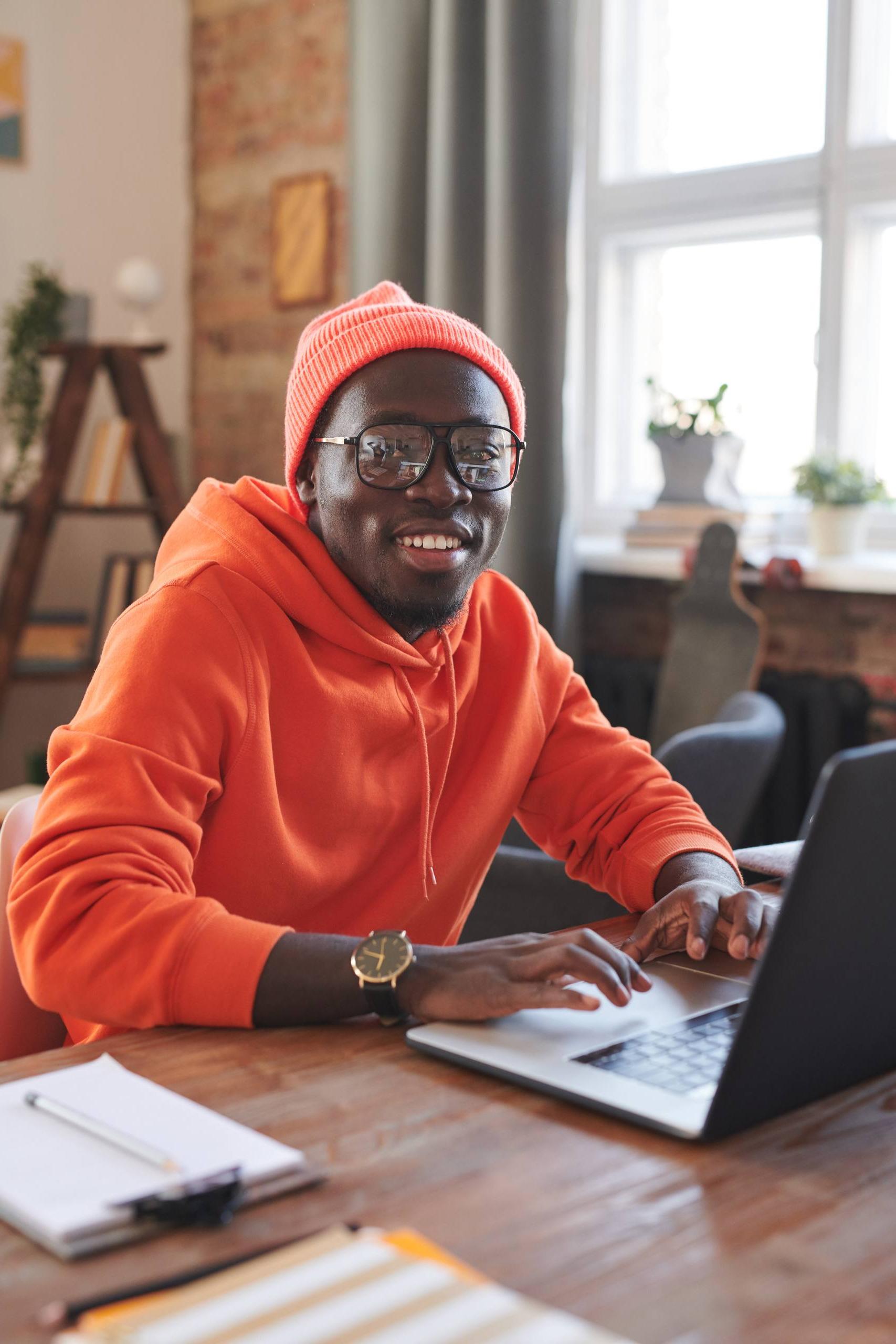 快乐的非洲裔美国人的垂直肖像，穿着鲜艳的橙色衣服，坐在家里的办公桌前，在笔记本电脑上上网，对着镜头微笑